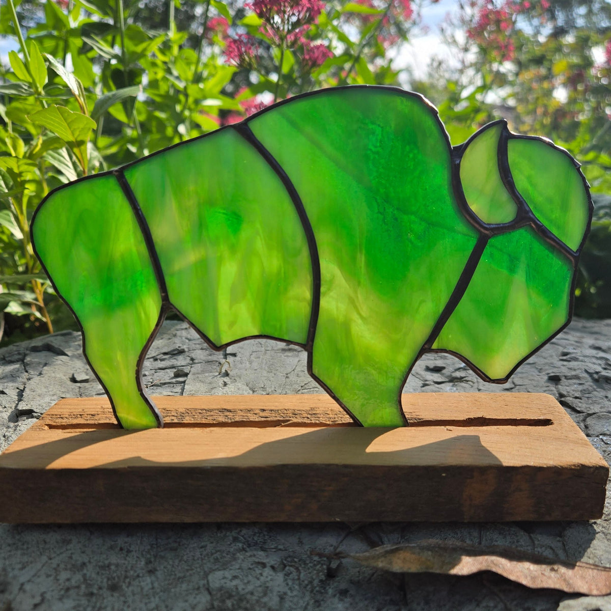 A green stained-glass-style bison silhouette mounted on a rectangular wooden base, photographed outdoors on a stone surface with blurred greenery and flowers in the background.